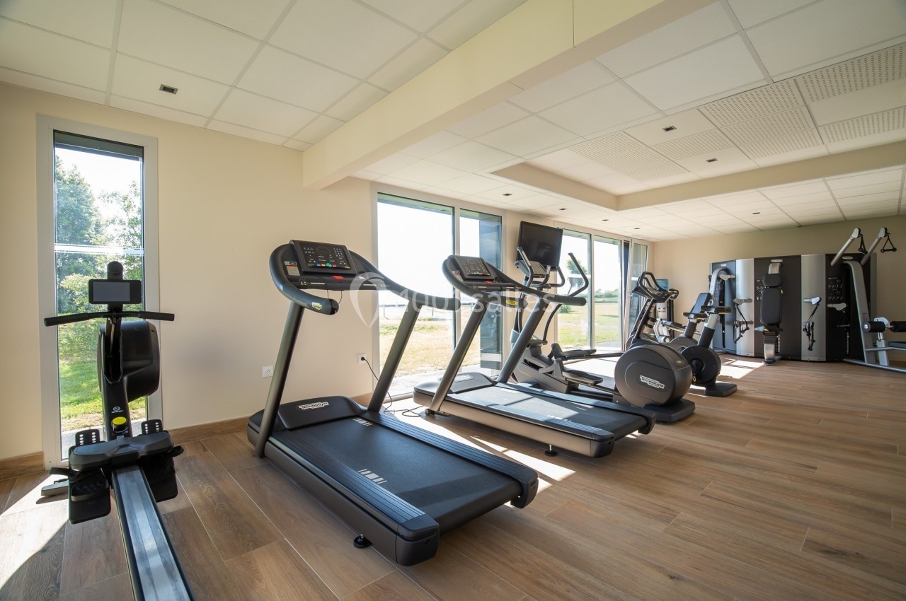 Salle de sport lumineuse avec tapis de course, rameur, vélos d'exercice et équipements de musculation modernes.