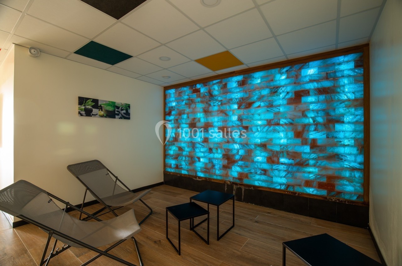 Mur lumineux bleu dans une salle de détente avec chaises longues, tables basses et plafond à dalles colorées.