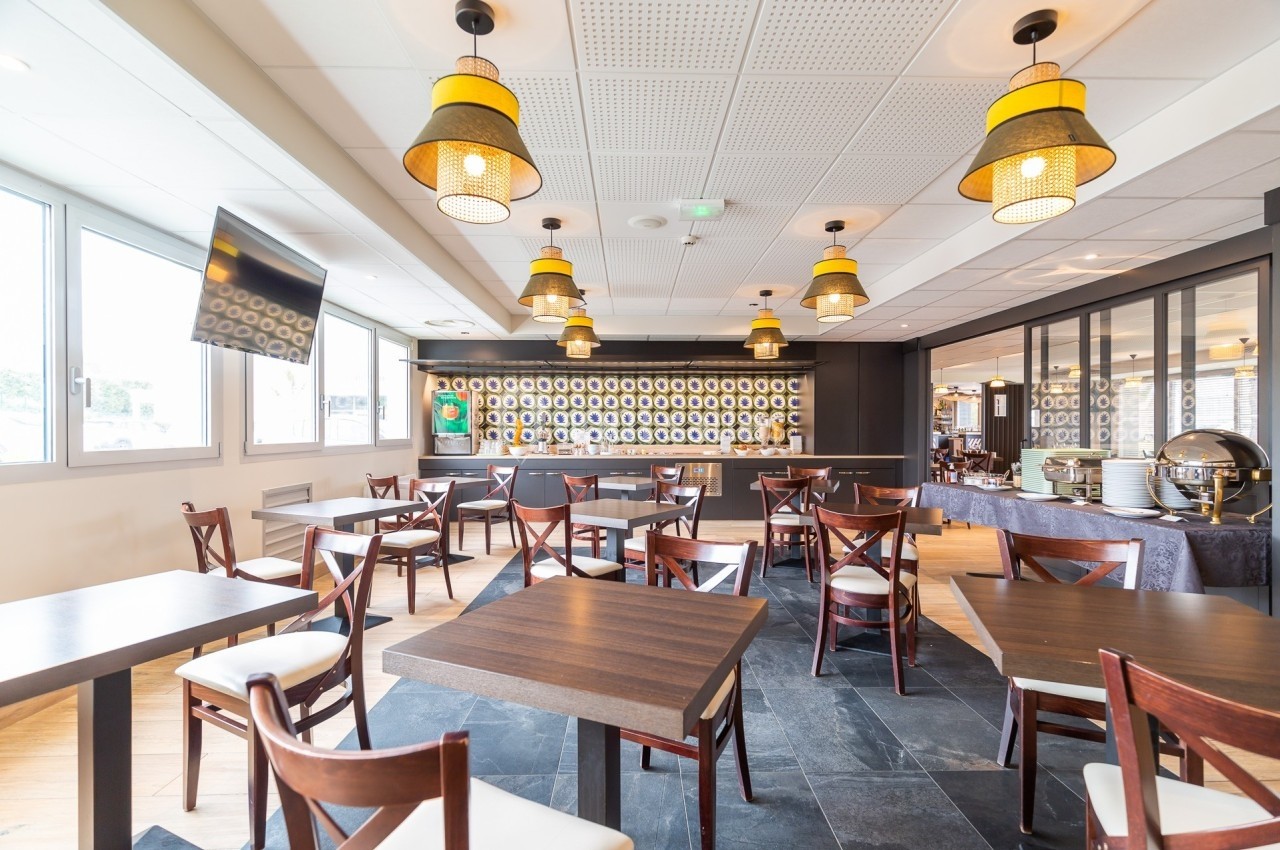 Salle de restaurant lumineuse avec tables en bois, chaises croisées et suspensions décoratives au plafond.