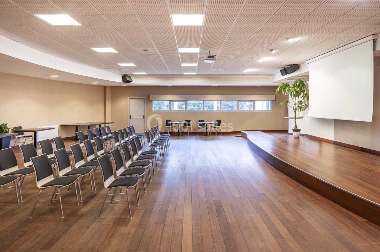 Salle de conférence lumineuse avec chaises alignées, scène en bois, écran de projection et grandes fenêtres.
