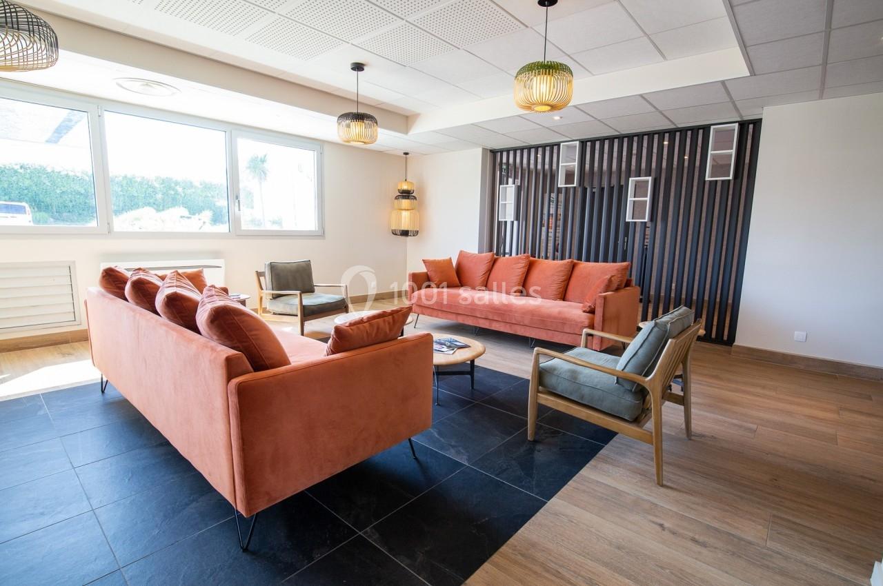 Salon lumineux avec deux canapés orange, fauteuils en bois, sol en carrelage et parquet, et grande fenêtre.