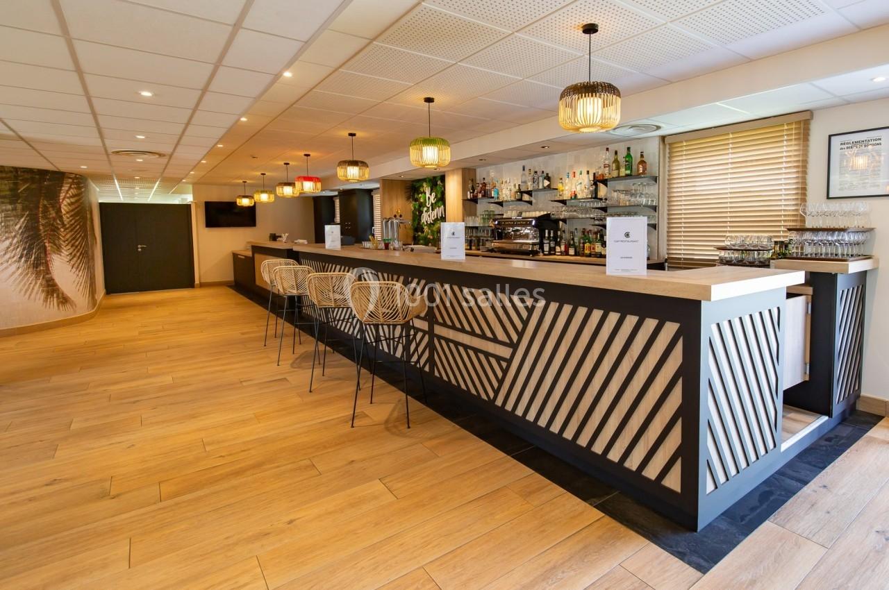 Bar moderne avec comptoir en bois, tabourets design, étagères de bouteilles et éclairage suspendu dans une salle lumineuse.