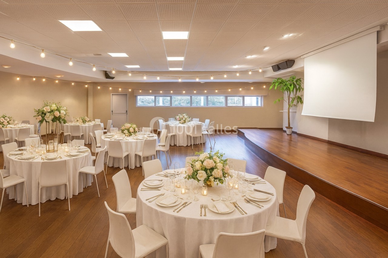 Salle de réception avec tables rondes décorées de fleurs et bougies, éclairage doux et scène en bois.