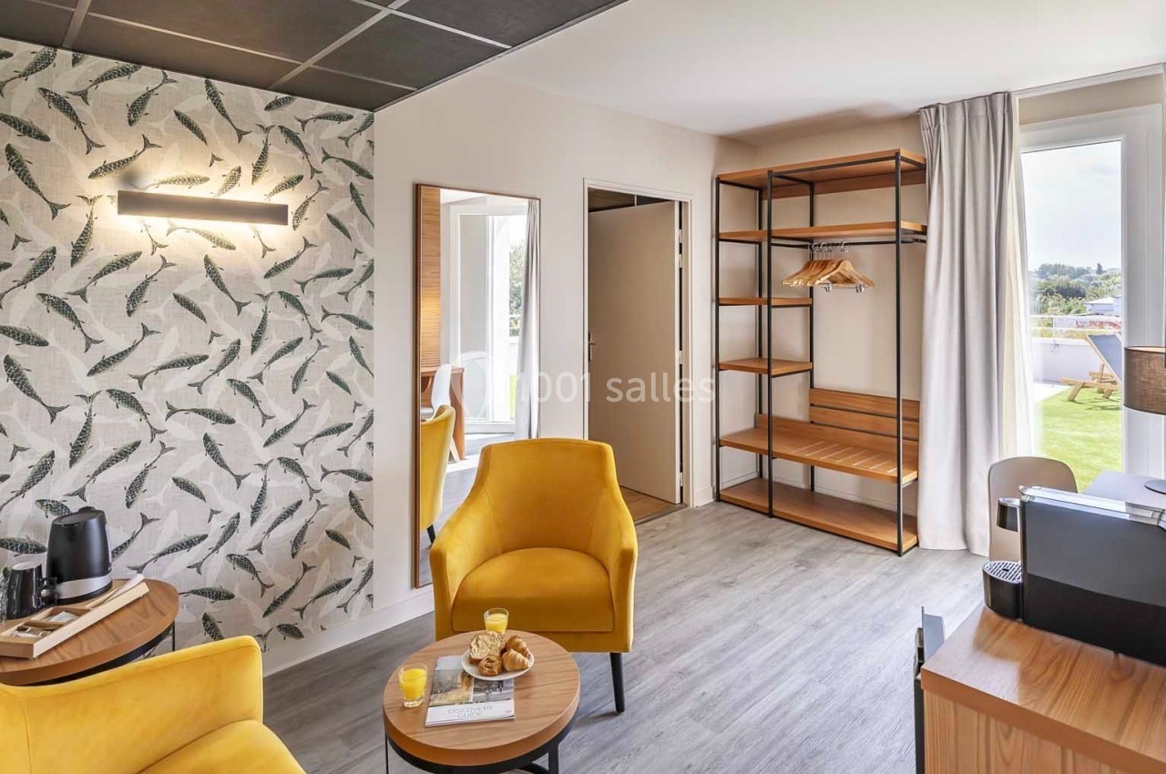 Espace lumineux avec fauteuils jaunes, table basse, étagères en bois, et vue sur une terrasse extérieure.