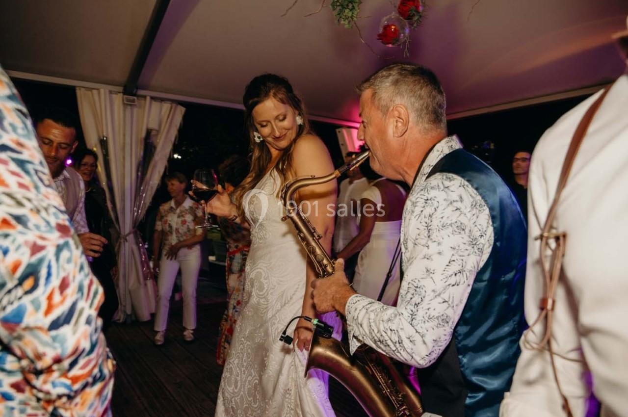Une femme en robe blanche danse avec un saxophoniste jouant lors d'une soirée festive en intérieur.