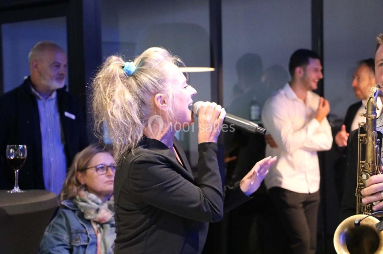 Une chanteuse interprète un morceau au micro lors d'un événement, entourée de spectateurs en arrière-plan.