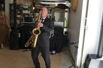 Un musicien en costume joue du saxophone dans un espace intérieur décoré avec des étagères et des bouteilles.