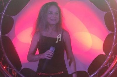 Femme tenant un micro, debout devant un décor lumineux rose et rouge avec une structure circulaire en arrière-plan.