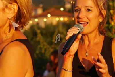 Une femme chante au micro en extérieur, sous une lumière dorée, avec un décor flou de soirée en arrière-plan.
