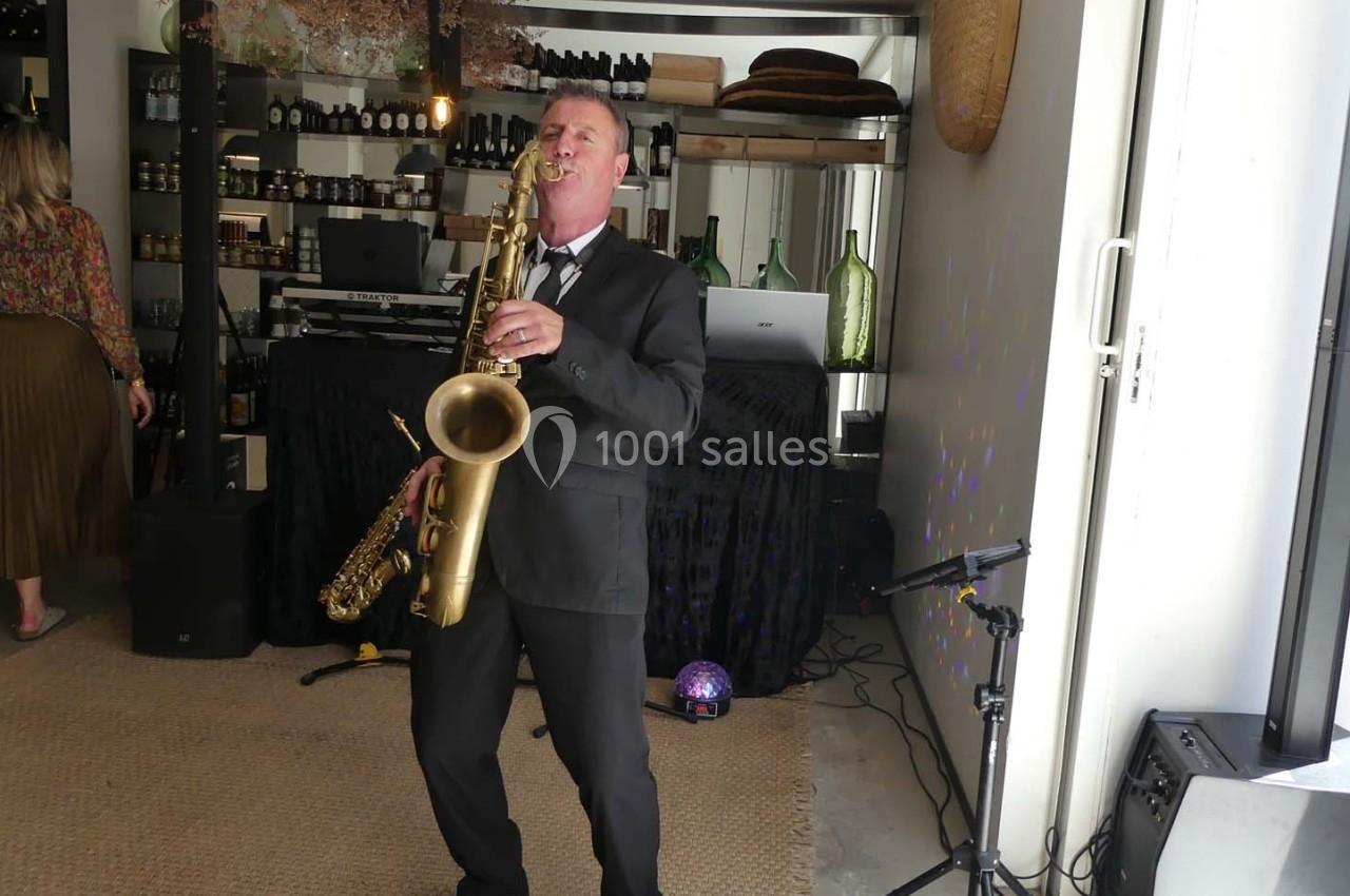Un musicien en costume joue du saxophone dans un espace intérieur décoré avec des étagères et des bouteilles.