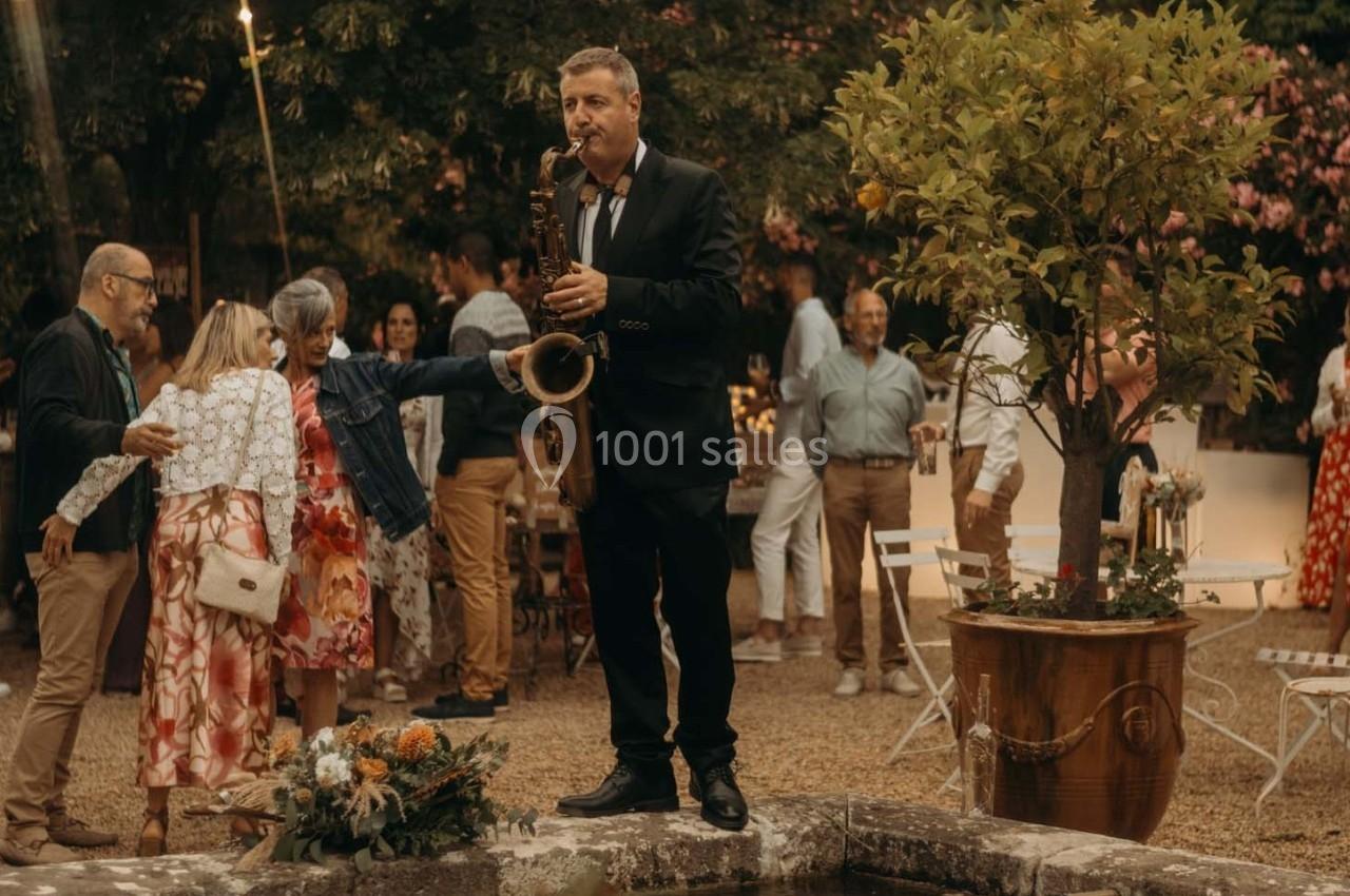 Un musicien joue du saxophone debout sur un rebord en pierre lors d'une fête en plein air avec des invités dansant.