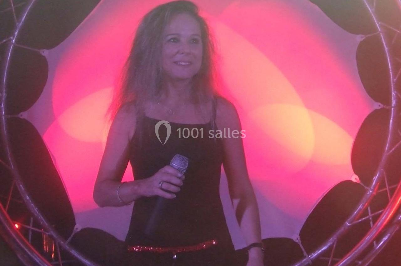 Femme tenant un micro, debout devant un fond lumineux rose et rouge avec une structure circulaire métallique.