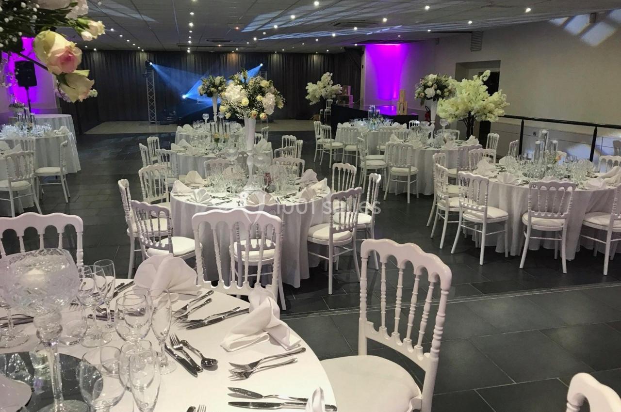 Salle de réception élégamment décorée avec des tables rondes, nappes blanches, chaises assorties et éclairage coloré.