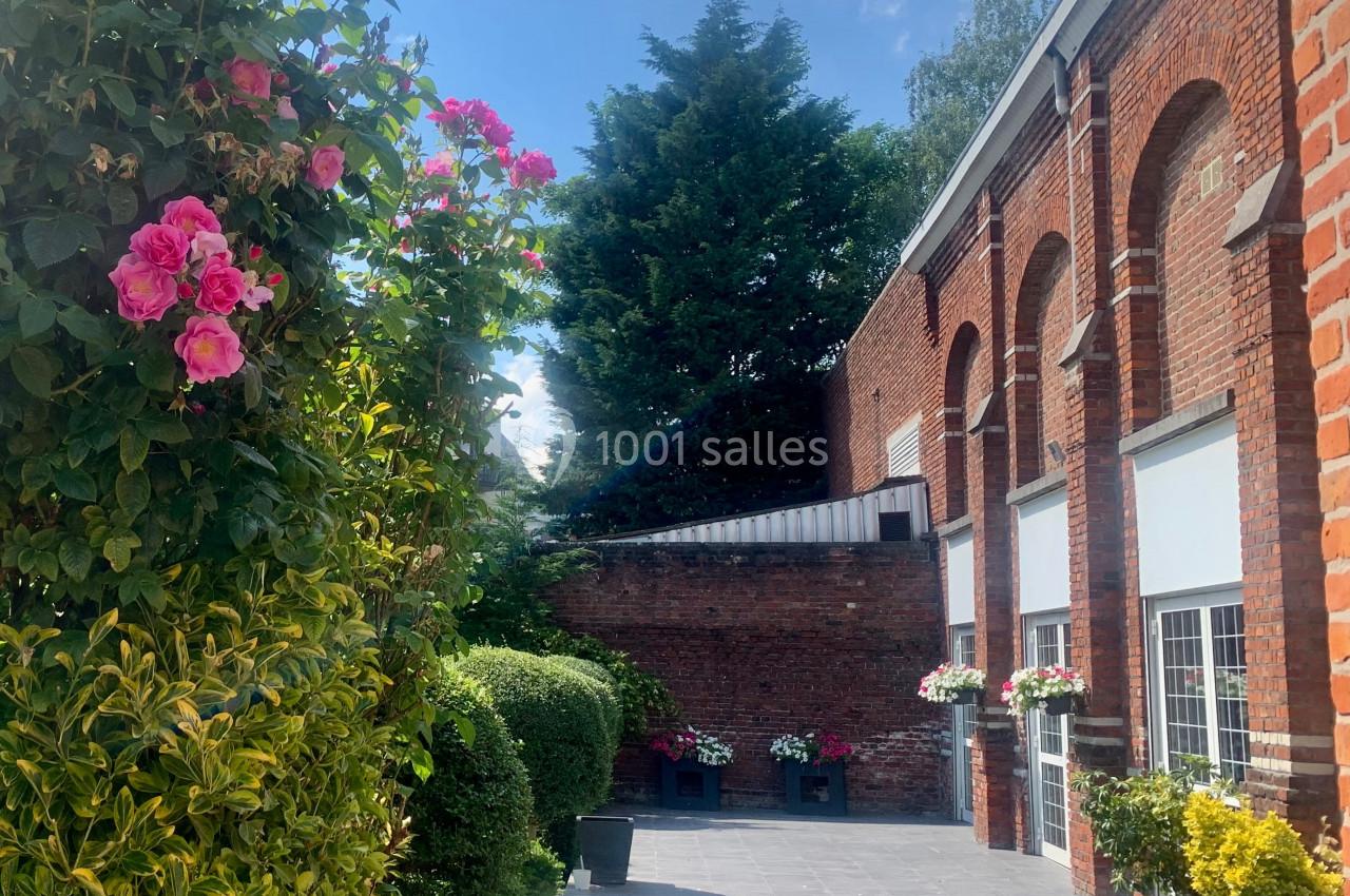 Allée pavée bordée de rosiers et de buissons, longeant un bâtiment en briques sous un ciel ensoleillé.
