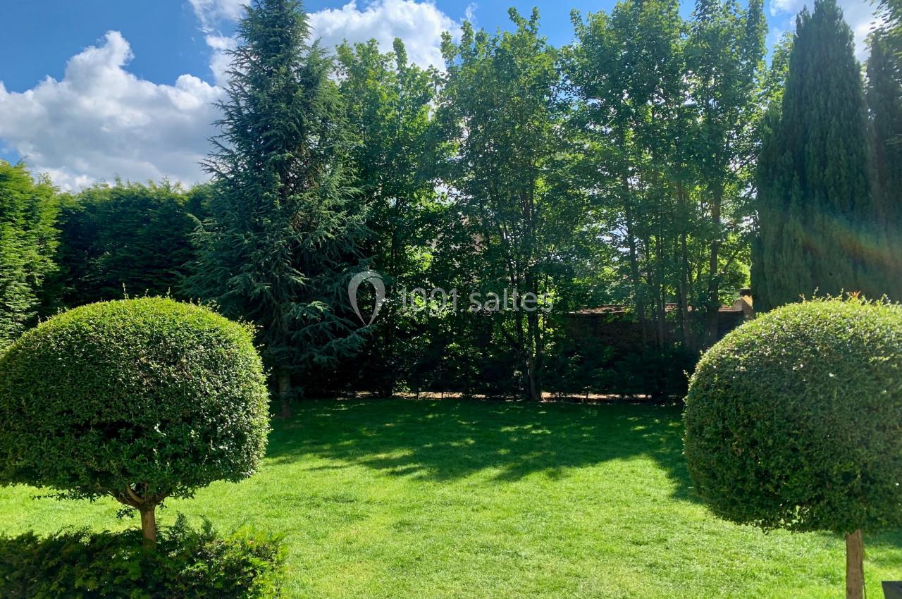 Jardin verdoyant avec pelouse, arbres et arbustes taillés, sous un ciel bleu parsemé de nuages.