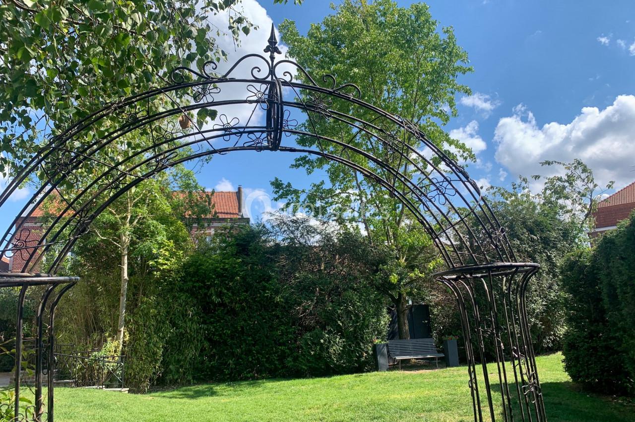 Arche en fer forgé dans un jardin verdoyant avec pelouse, arbres et ciel bleu parsemé de nuages.