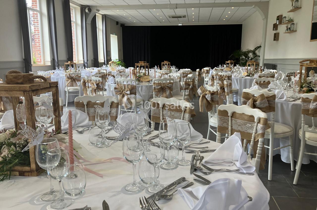 Salle de réception décorée pour un mariage, avec tables dressées, nappes blanches et chaises ornées de rubans beiges.