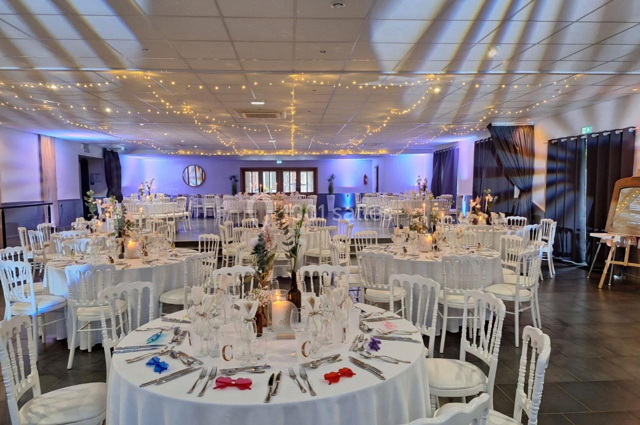 Salle de réception décorée avec des tables rondes dressées, chaises blanches et guirlandes lumineuses suspendues.