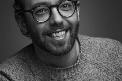 Femme assise, vêtue d'une robe blanche fluide, posant avec une expression sereine sur fond neutre.