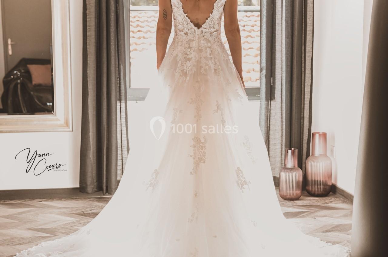 Une femme en robe de mariée vue de dos, debout devant une fenêtre laissant entrer la lumière naturelle.