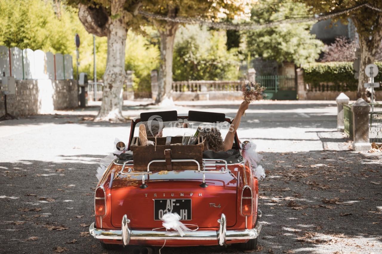 Deux personnes dans une voiture décapotable rouge, l'une tenant un bouquet, sur une route bordée d'arbres.