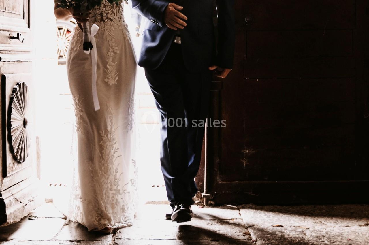 Une mariée en robe blanche entre dans un bâtiment sombre, accompagnée d'un homme en costume tenant son bras.