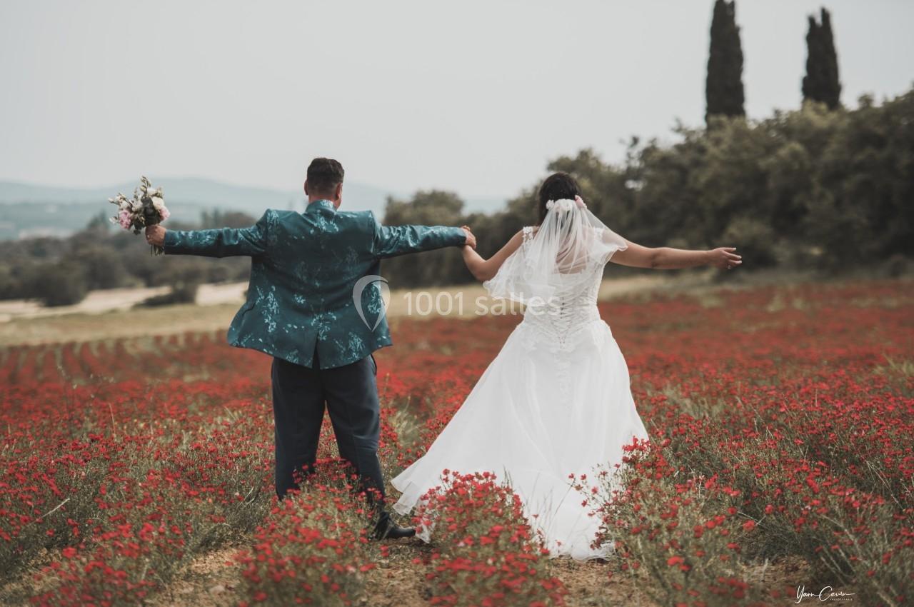 Un couple de mariés de dos, bras écartés, debout dans un champ de fleurs rouges avec des arbres en arrière-plan.