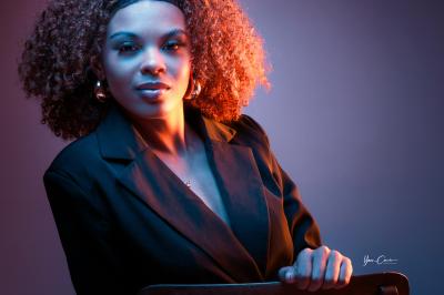 Femme assise, vêtue d'une robe blanche fluide, posant avec une expression sereine sur fond neutre.