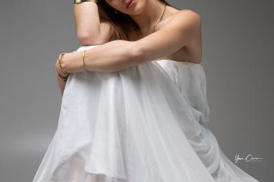 Femme assise, vêtue d'une robe blanche fluide, posant avec une expression sereine sur fond neutre.