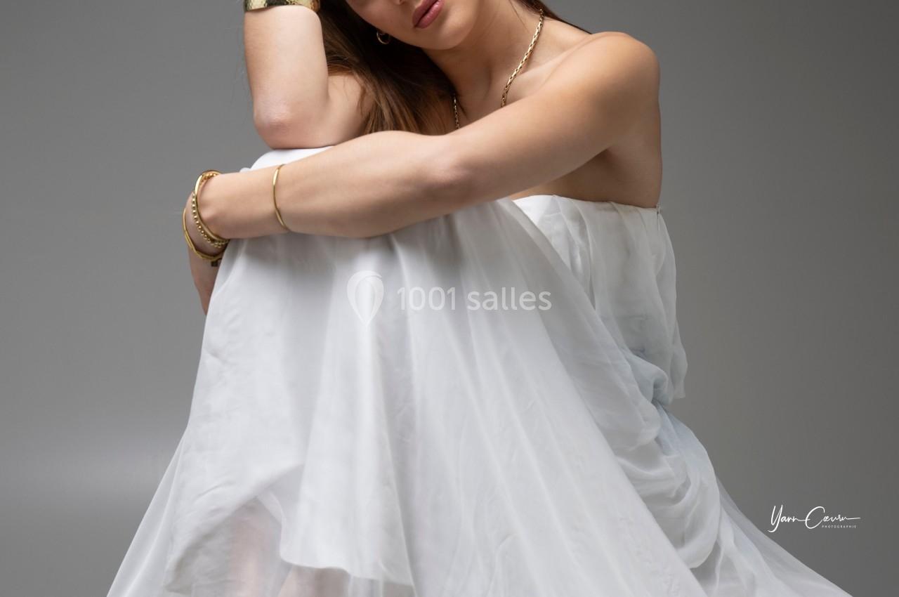 Femme assise, vêtue d'une robe blanche fluide, posant avec une expression sereine sur fond neutre.