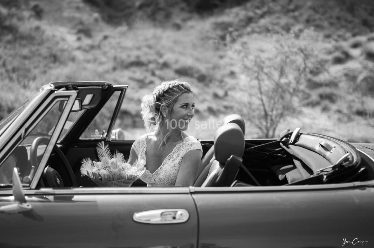 Une femme en robe de mariée sourit assise dans une voiture décapotable, tenant un bouquet de fleurs.