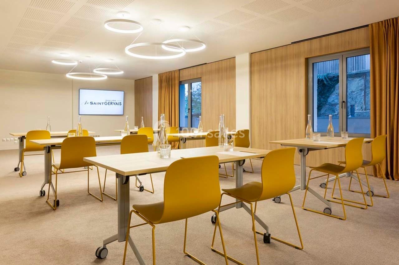 Salle de réunion lumineuse avec tables et chaises jaunes, écran mural et grandes fenêtres avec rideaux.