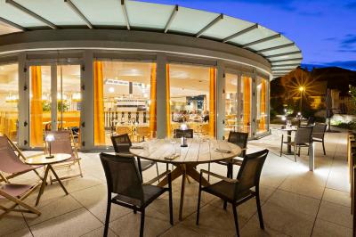 Façade d'un bâtiment éclairé la nuit, entouré d'un jardin avec des arbres, des chaises et des tables en extérieur.