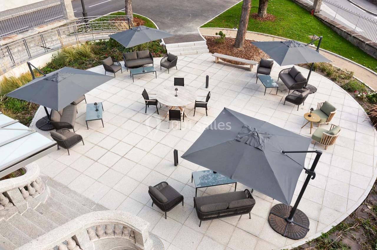 Terrasse aménagée avec des tables, chaises, canapés et parasols gris sur un sol en dalles claires.
