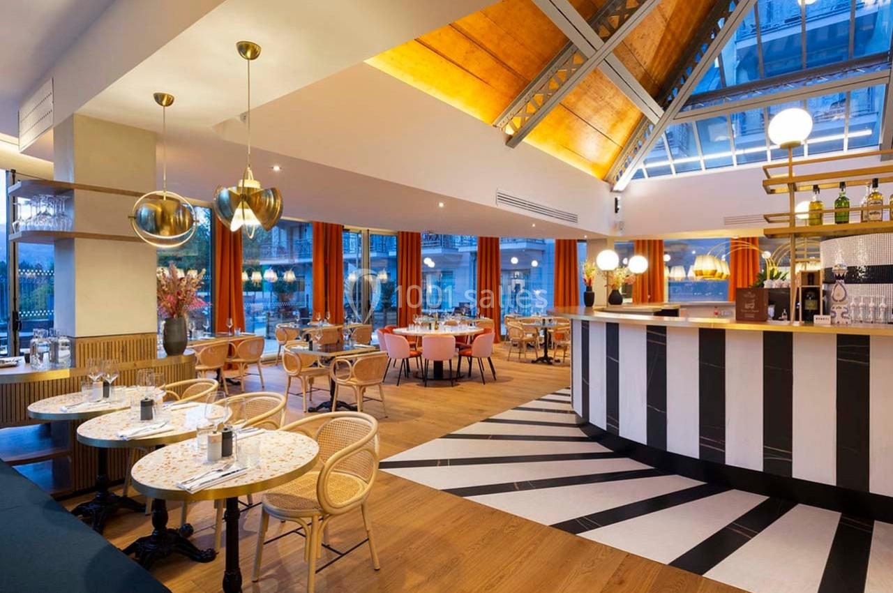 Salle de restaurant lumineuse avec mobilier en bois, tables dressées, baies vitrées et plafond en verrière.