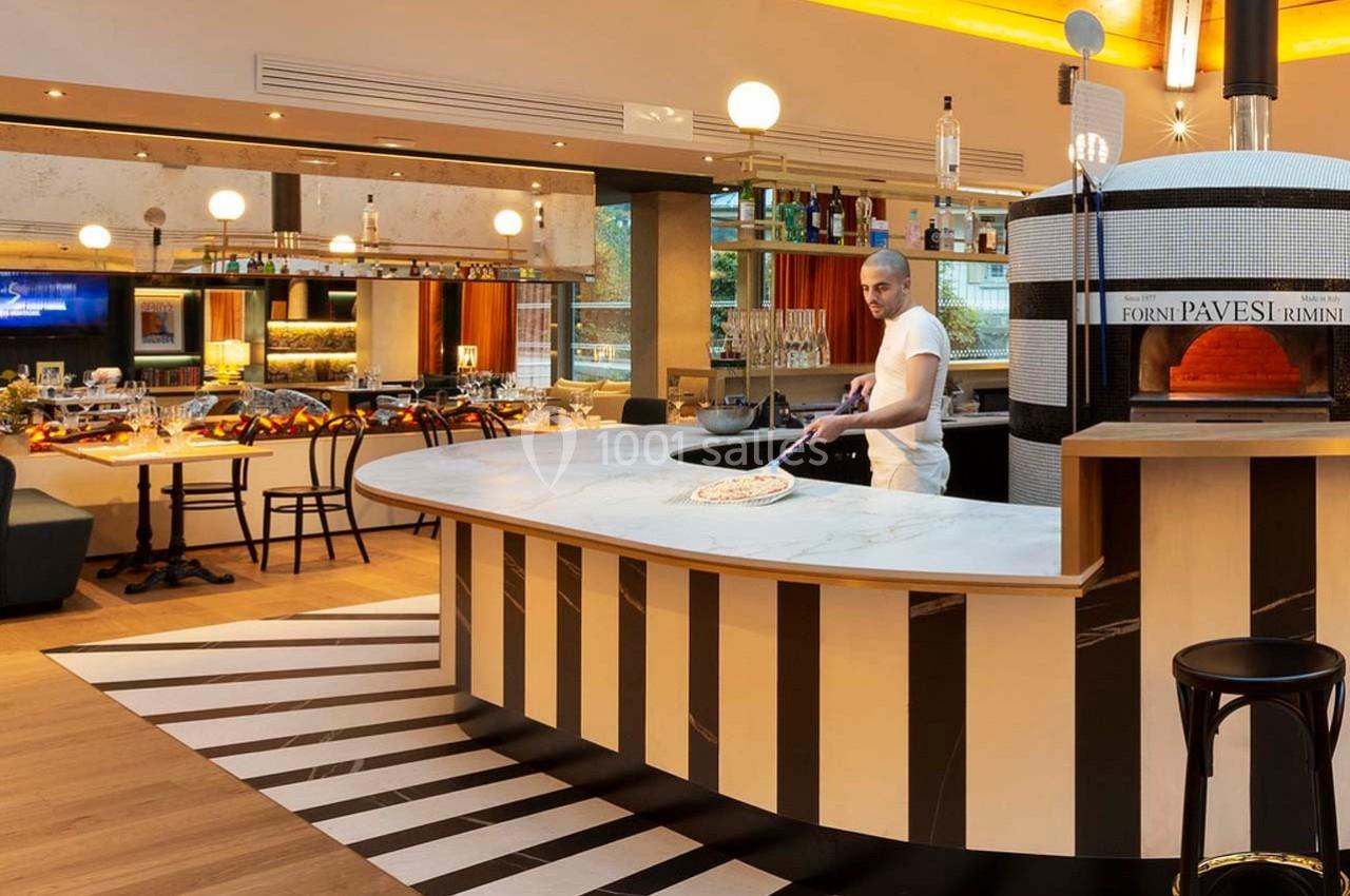 Un pizzaiolo prépare une pizza devant un four à bois dans un restaurant au décor moderne et chaleureux.