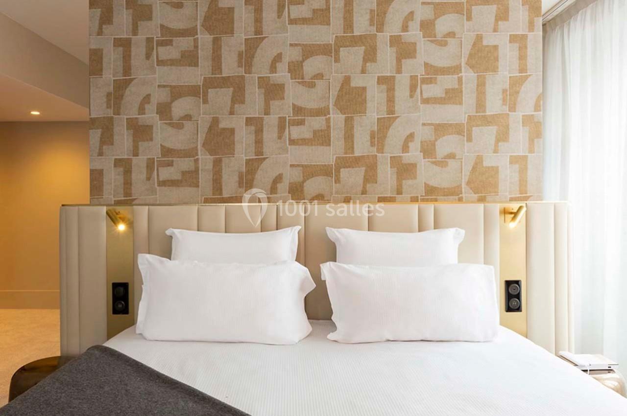 Lit double avec tête de lit beige capitonnée, draps blancs et mur décoratif à motifs géométriques en arrière-plan.