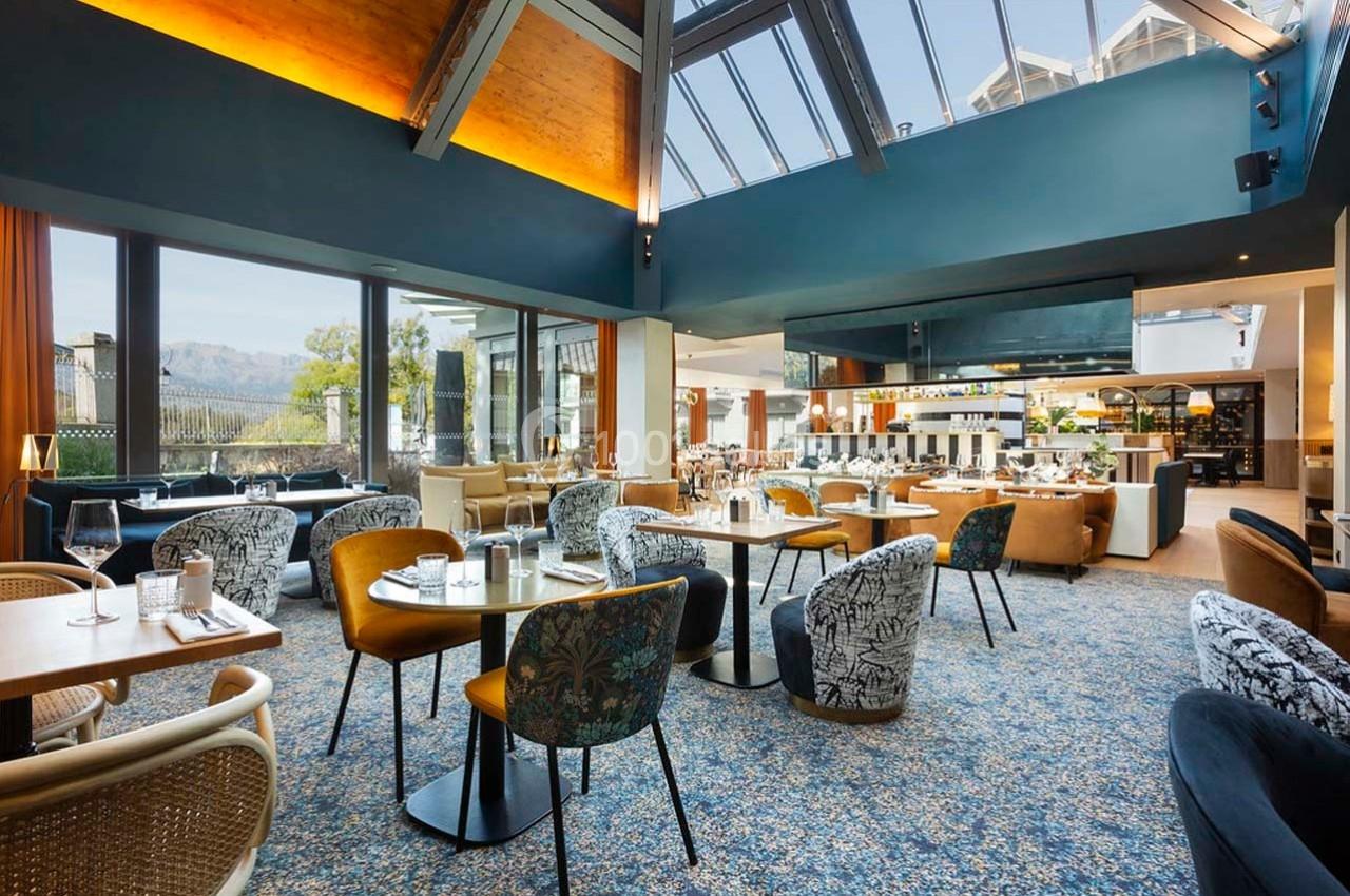 Salle de restaurant lumineuse avec de grandes baies vitrées, mobilier varié et décoration moderne.