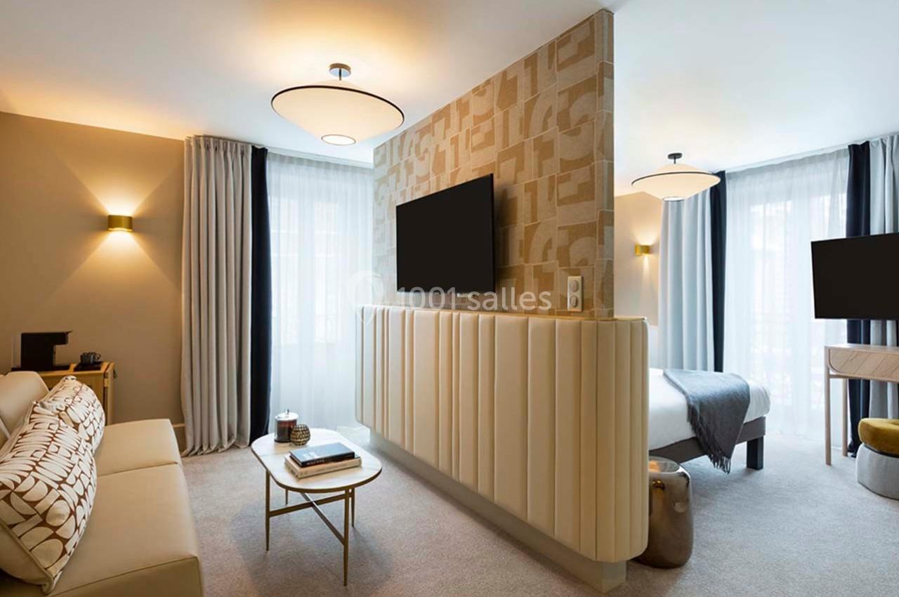 Salon et chambre d'hôtel modernes séparés par une cloison, avec mobilier élégant et éclairage doux.