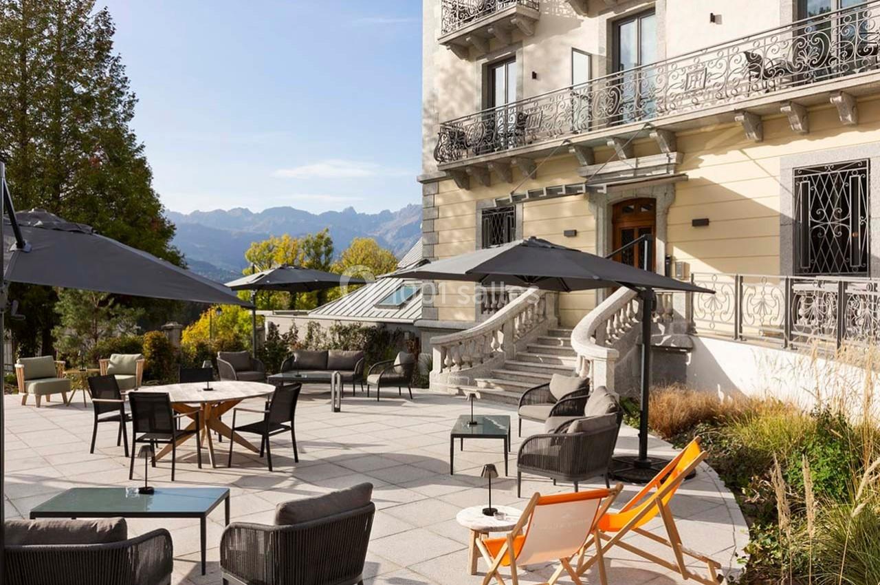 Terrasse aménagée avec tables, chaises et parasols, devant un bâtiment élégant, entourée de végétation et montagnes.