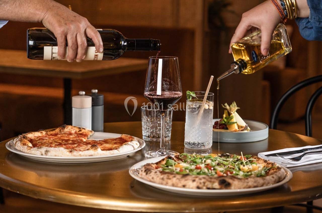 Deux personnes servent du vin rouge et un cocktail à une table avec des pizzas et des condiments.