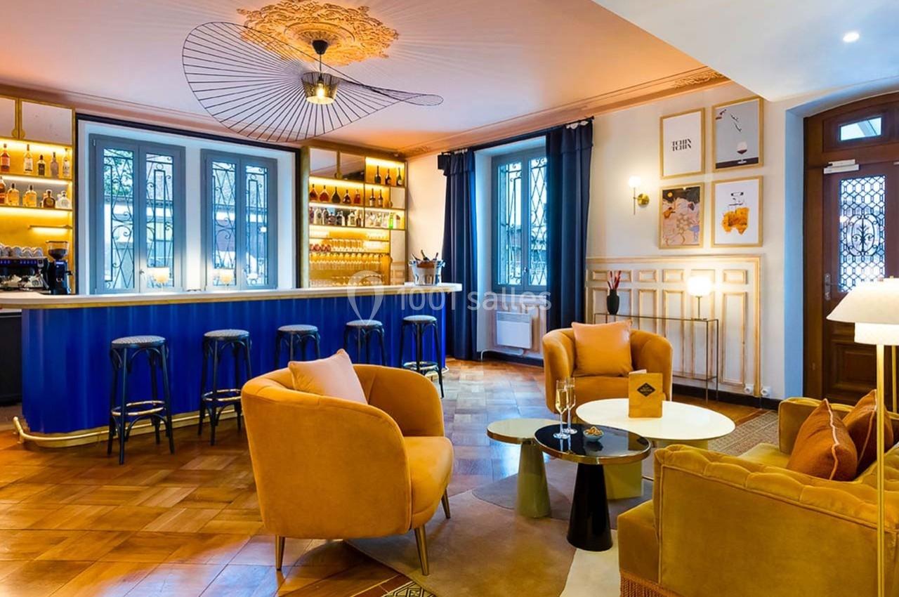 Salon élégant avec bar bleu, fauteuils jaunes, tables basses et éclairage chaleureux dans un décor raffiné.