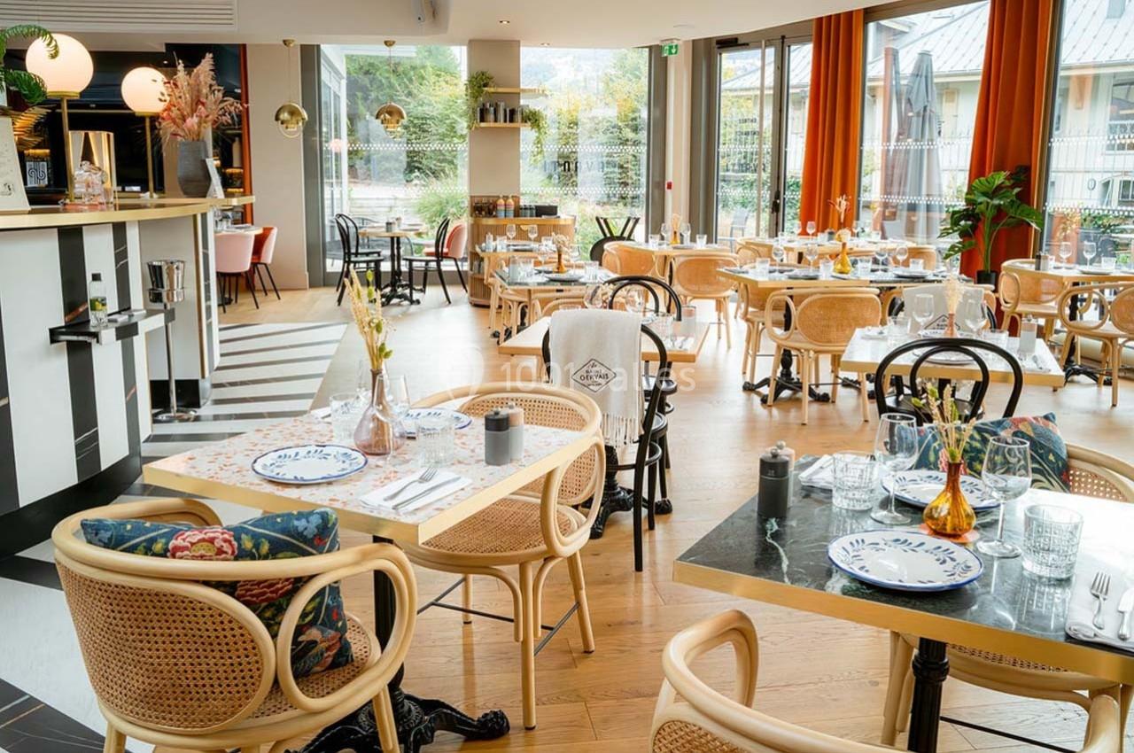 Salle de restaurant lumineuse avec tables dressées, chaises en rotin et grandes baies vitrées donnant sur un jardin.