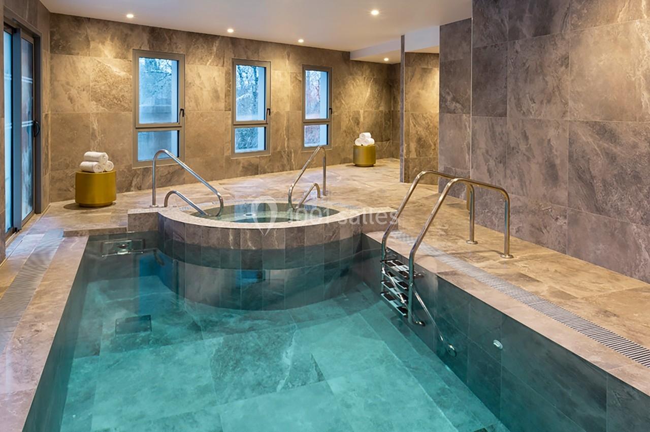 Piscine intérieure avec jacuzzi intégré, entourée de murs en pierre grise et éclairée par des fenêtres et des spots au…