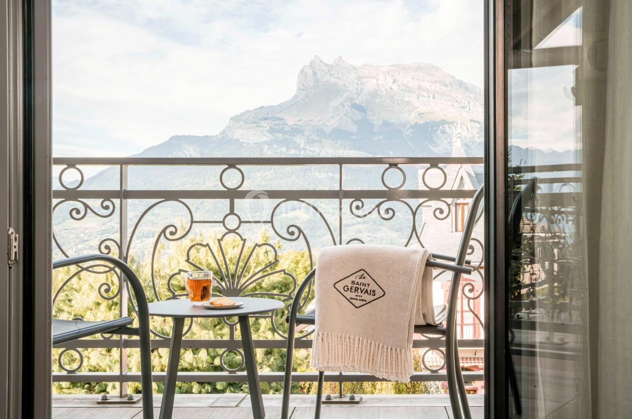 Vue d'un balcon avec table, tasse de thé et plaid, donnant sur une montagne sous un ciel partiellement nuageux.