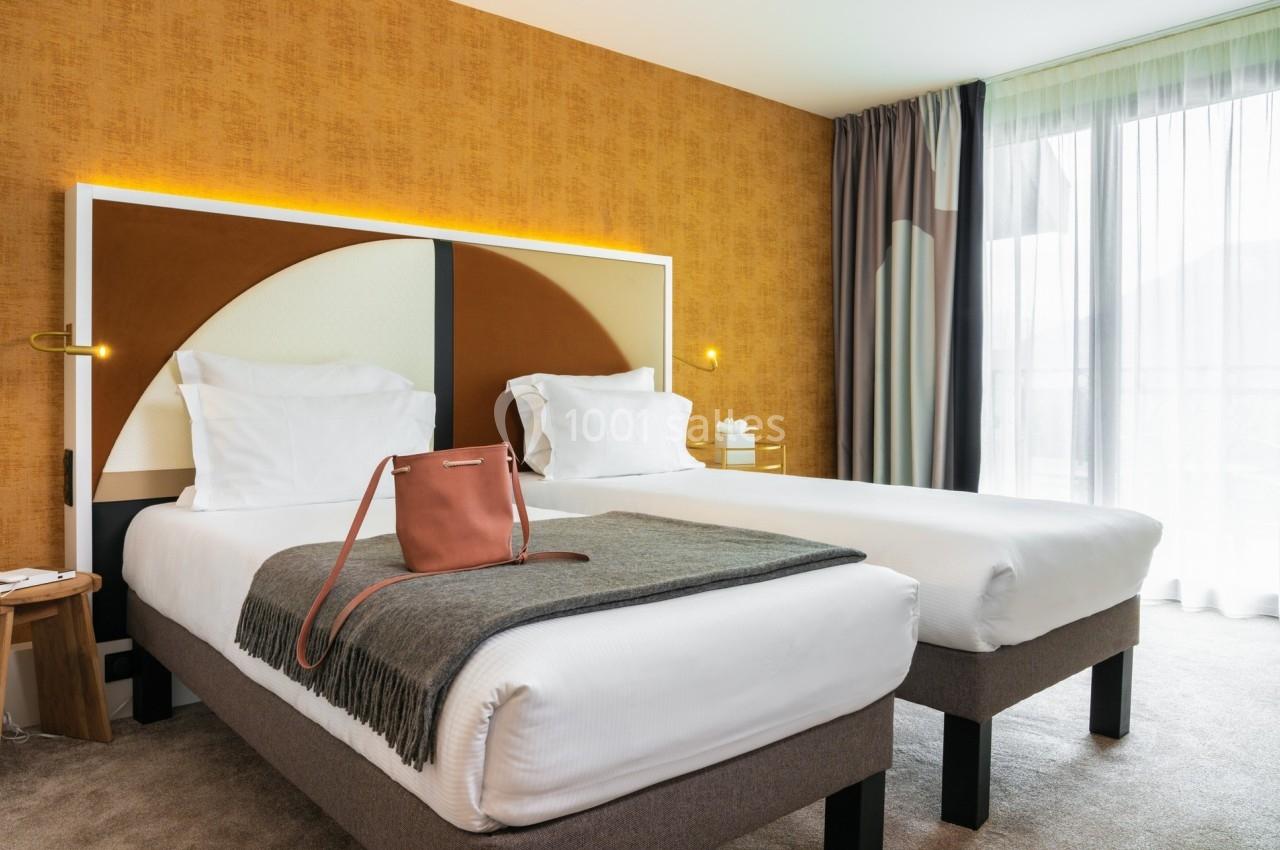 Deux lits simples avec tête de lit moderne, plaid gris et sac posé, dans une chambre lumineuse aux tons chauds.