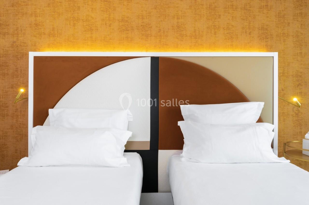 Deux lits simples avec draps blancs, tête de lit géométrique et mur texturé orange en arrière-plan.