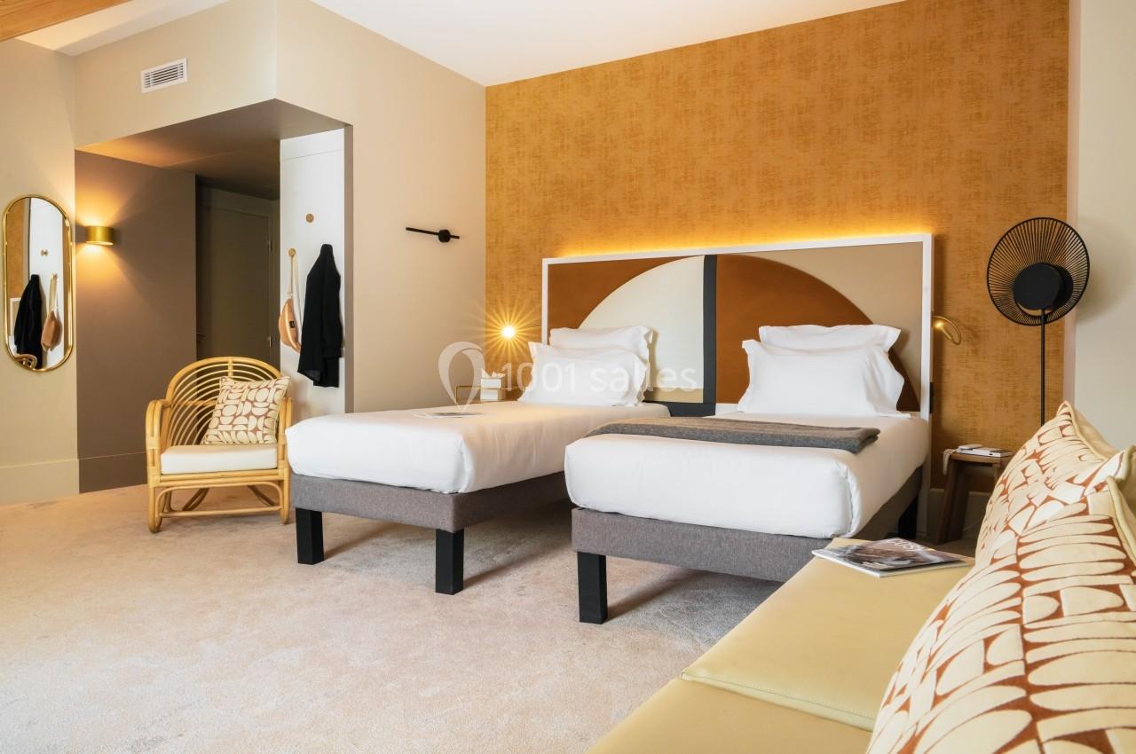 Chambre d'hôtel lumineuse avec deux lits simples, fauteuil en rotin, mur ocre et décoration moderne.