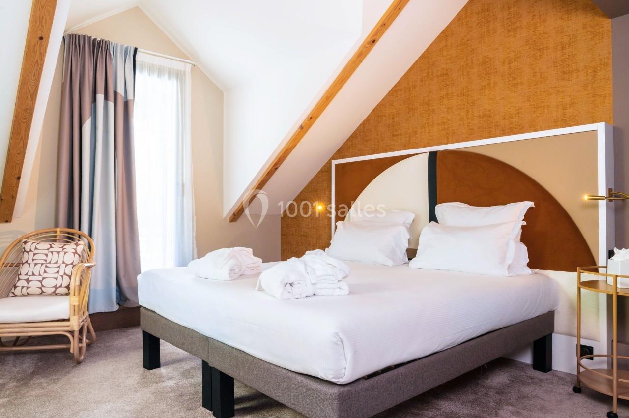 Chambre lumineuse sous combles avec lit double, fauteuil en rotin, rideaux épais et décoration murale moderne.