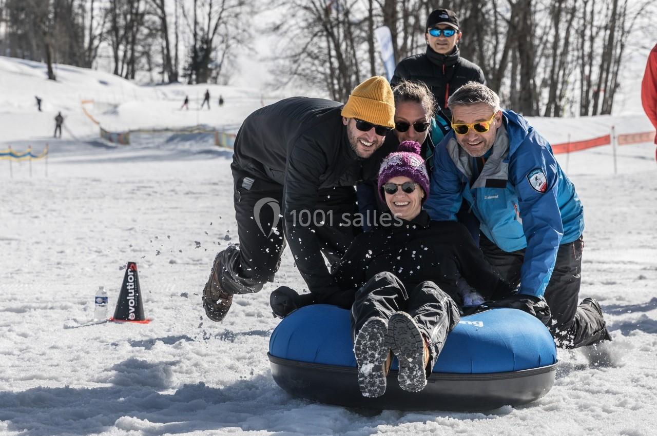 Des personnes glissent sur une bouée dans la neige, entourées d'autres participants et d'arbres en arrière-plan.